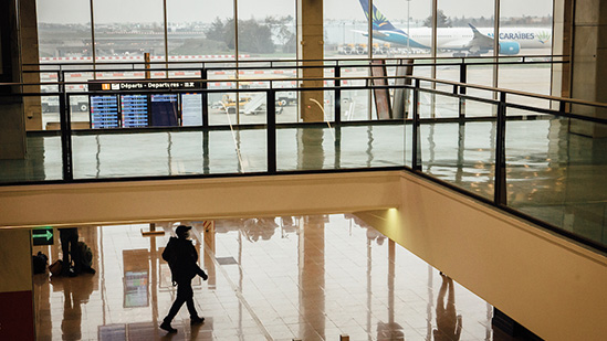 A silhouette of a traveler with a backpack walking through an airport terminal. The scene captures reflections on the polished floor, a departures board, and large windows revealing a parked airplane on the tarmac outside.