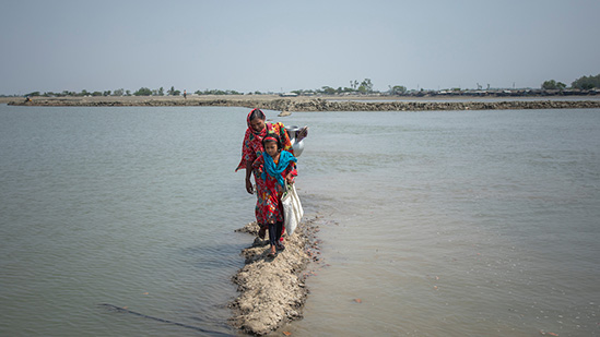 A woman and a young girl walk along a narrow, muddy path surrounded by water. The woman is carrying a metal container on her head, and both are dressed in bright, patterned clothes. The background shows a distant shoreline under a clear sky.